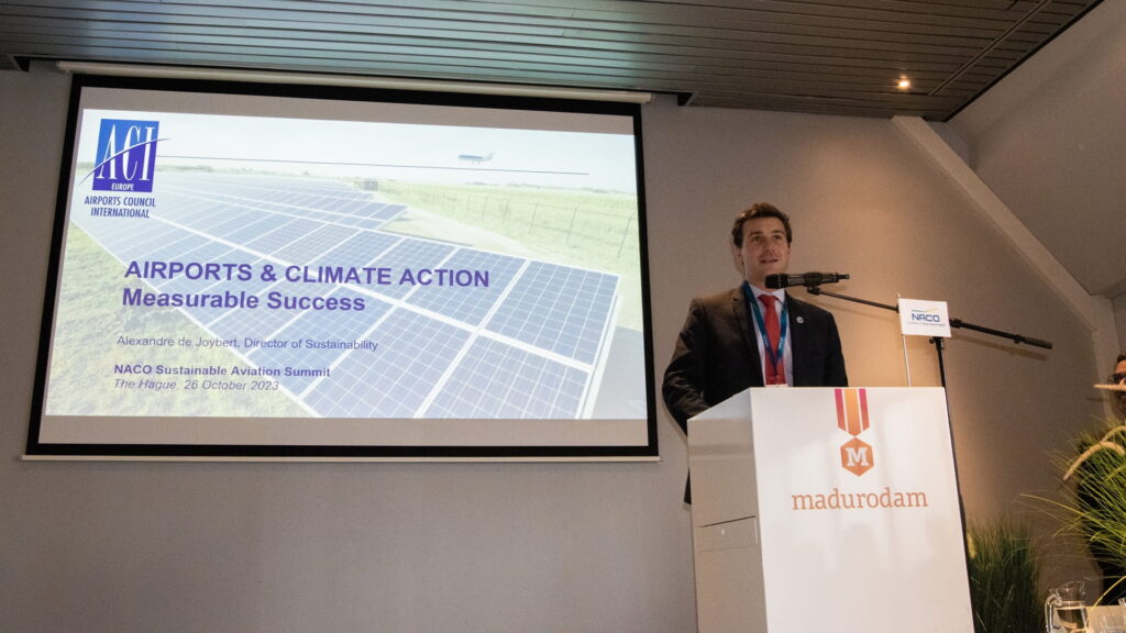 A speaker at a podium gives a presentation next to a projection. The projection shows solar panels at an airport