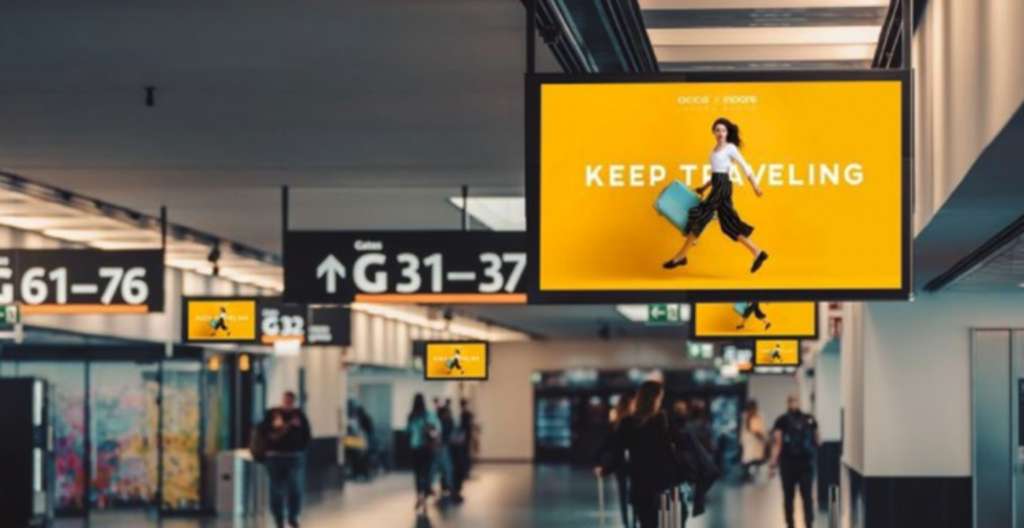 The inside of an airport terminal. The camera is focused on the ceiling where several displays show a yellow background with white text that reads "Keep Traveling". There is a woman walking with a suitcase in front of the text.