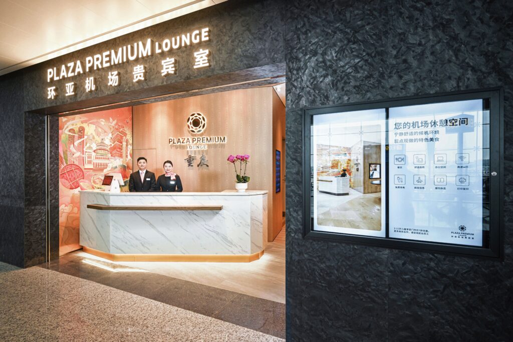 There is a black archway with the words "Plaza Premium Lounge" written over it. Past the archway, there's a white marble check in desk with two smiling members of staff stood behind it
