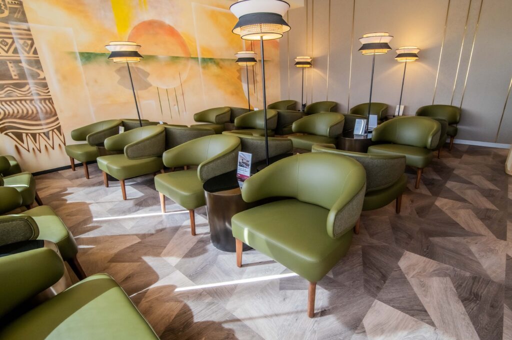 A light room is filled with forest green armchairs. Between the chairs there are woven standing lamps. In the background there is a mural of a sunrise.