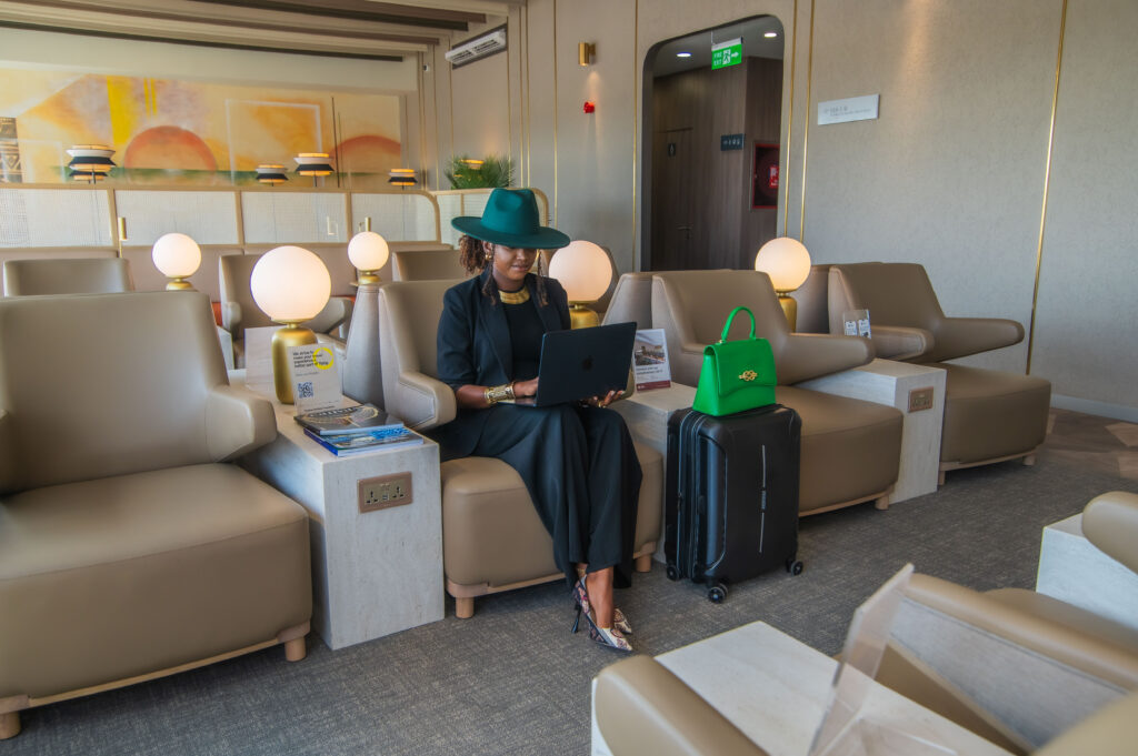 A woman sits on a light beige comfortable seat with her laptop on her lap, suitcases beside her and a teal hat on her head. The rest of the room is light, warm and inviting - packed with identical beige seats