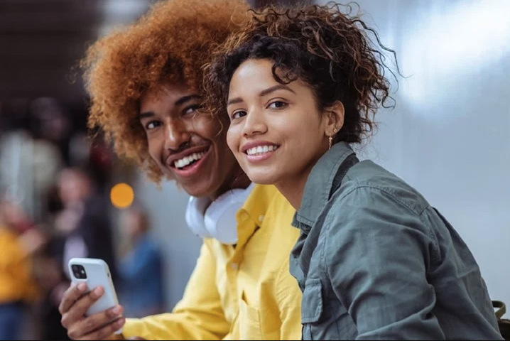 A woman in a green coat and a man in a yellow coat holding a phone both smiling at the camera