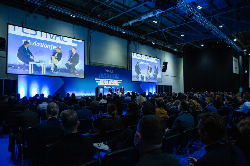 A photo from the crowd of a panel discussion at World Aviation Festival 2023. The room is dark, 2 screens either side of the stage show the participants