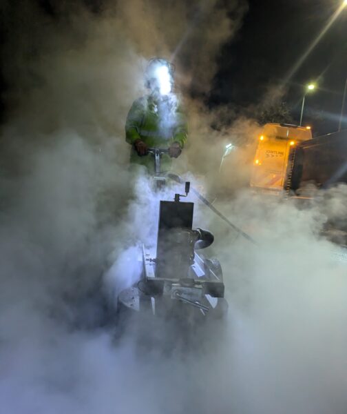 A worker operating a hydroblasting machine at night, surrounded by smoke