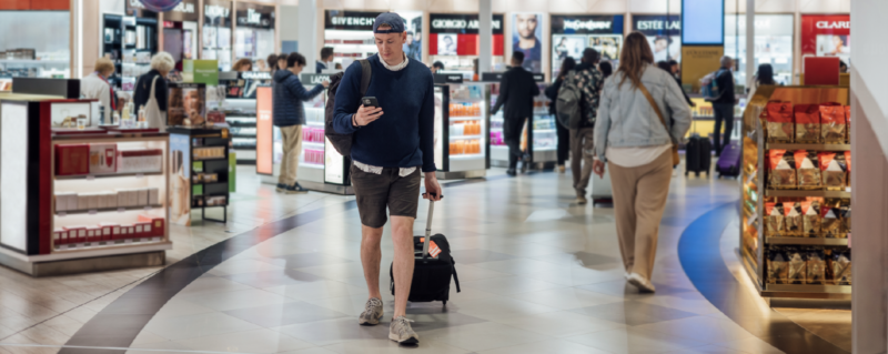 A man walking through airport duty free, pulling a wheeled suitcase in one hand and holding his phone with the other hand