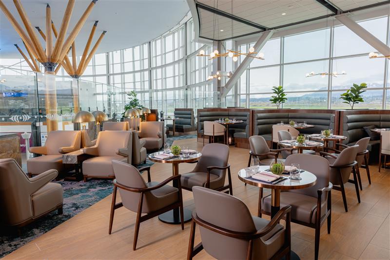 An airport lounge with round wooden tables and chairs with beige furnishings