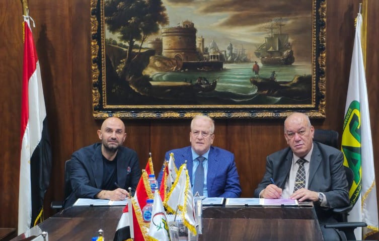 3 men from the respective companies, sat at a large brown desk with flags either side, signing the paper agreements