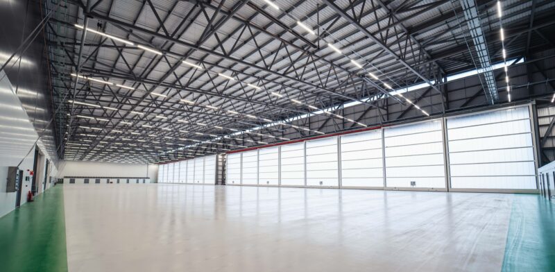 Interior of an aircraft hangar at an airport, open empty space with white flooring and black beams across the roof