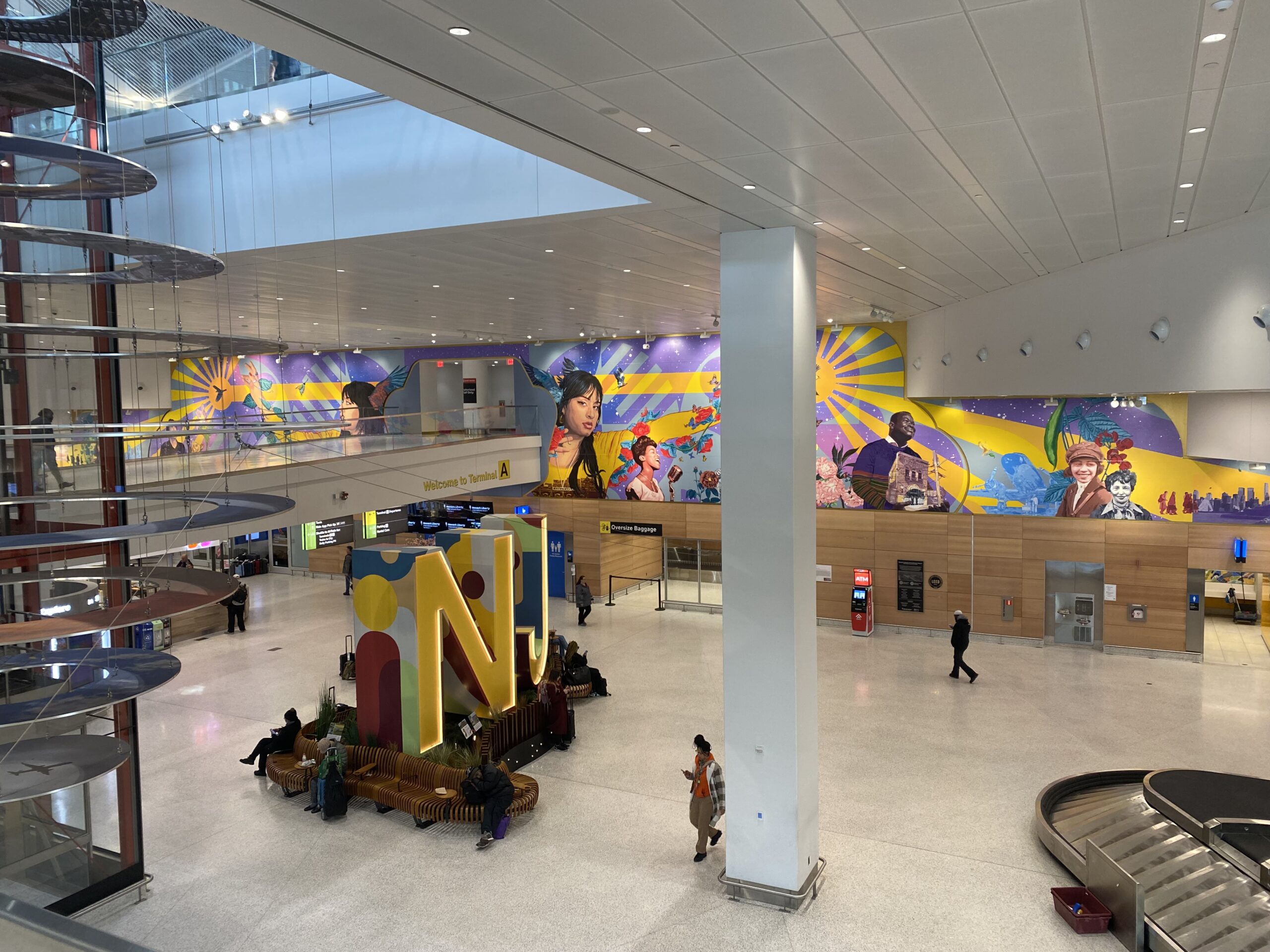 An expansive, brightly lit airport terminal features a large, colorful mural depicting diverse faces and scenes along a back wall, a prominent "N" sculpture in the center with seating, and various travelers and airport infrastructure.