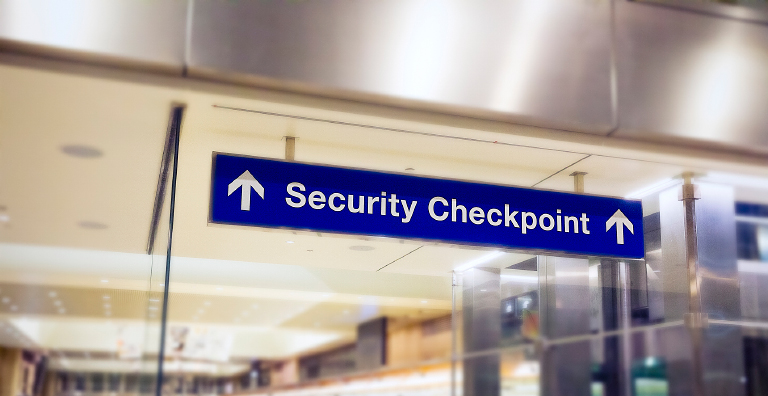 A blue rectangular sign with white text and arrows pointing right and left reads "Security Checkpoint" and hangs from the ceiling in an airport terminal.