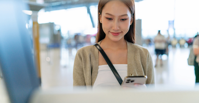 A smiling young Asian woman with long brown hair, wearing a white top and a beige cardigan, looks down at her smartphone while standing in what appears to be an airport, with a blurred kiosk or counter in the foreground.