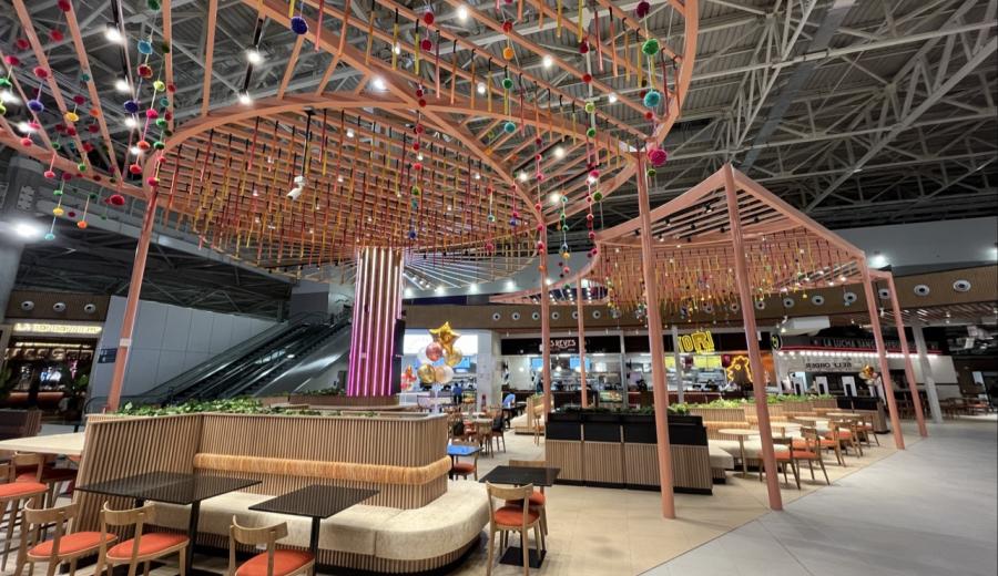 A vibrant food court features numerous small tables and banquette seating under an elaborate peach-colored canopy adorned with hanging colorful pom-poms and fabric strips.