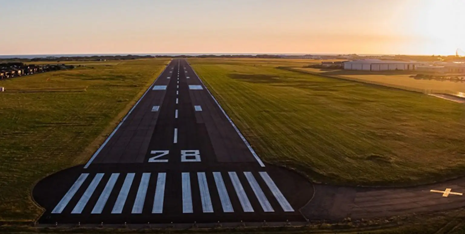An airport runway stretches into the distance, with green fields on either side and the ocean visible under a setting sun.