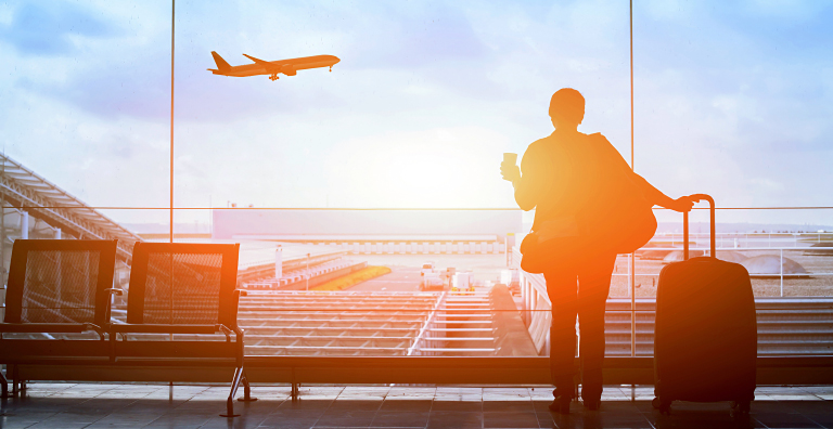 A silhouette of a traveler stands by a large airport window, holding a cup and pulling a rolling suitcase, looking out at the tarmac and a plane taking off, all bathed in bright sunlight.