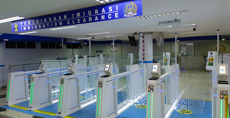 An array of modern immigration clearance gates with glowing green lights and digital displays are arranged in an airport or large building, under a prominent blue sign that reads "PEMERIKSAAN IMIGRASI IMMIGRATION CLEARANCE".