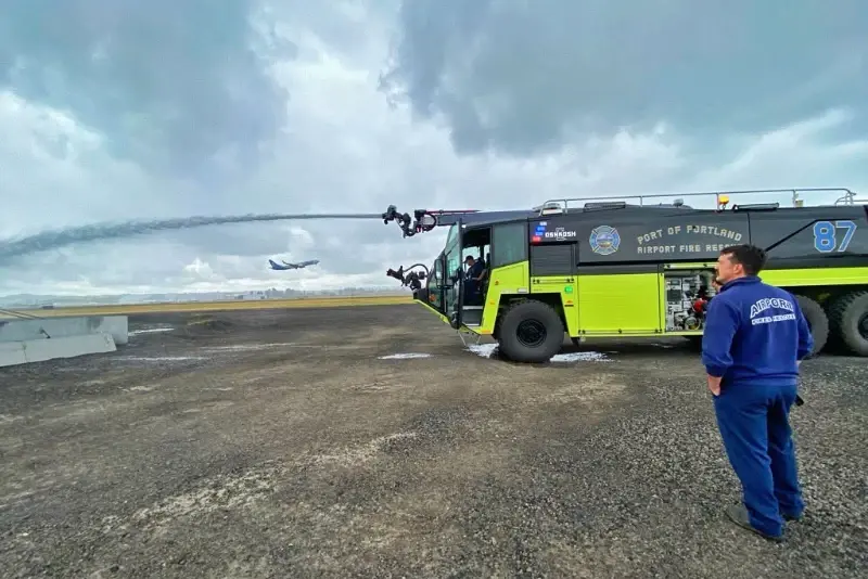 An ARFF firefighter is standing near a Striker 6x6 ARFF spraying water outside on a cloudy day