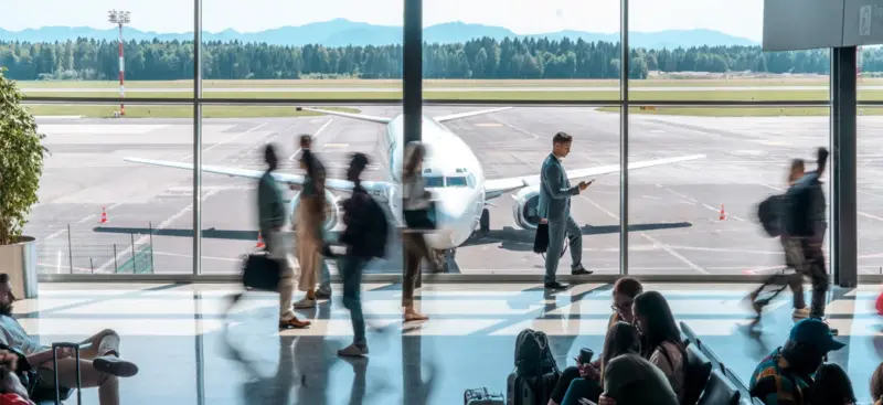 Passengers walking through an airport terminal past large windows overlooking a parked aircraft on the apron