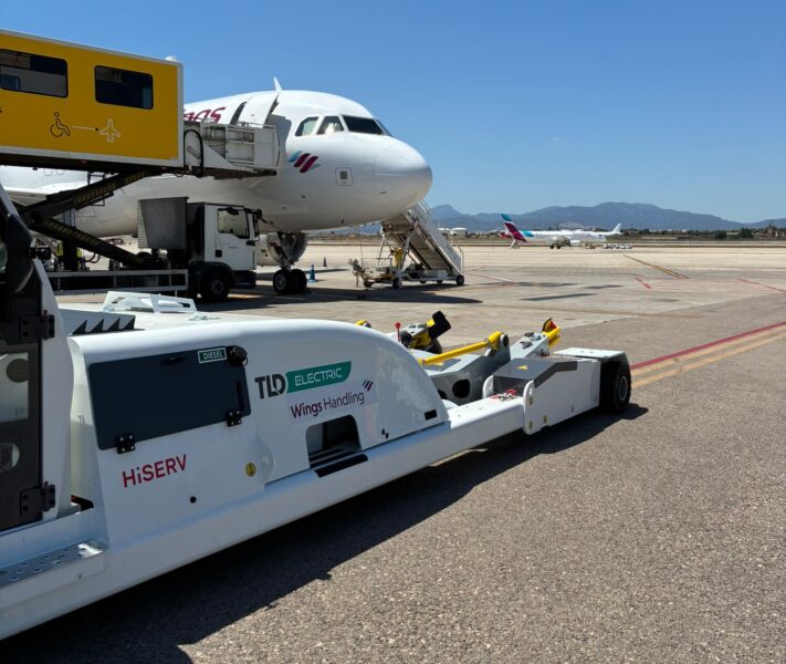 Electric towbarless aircraft tractor with HiSERV and Wings Handling branding on the apron beside a Eurowings aircraft and ambulift