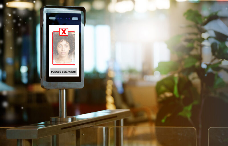Facial verification terminal mounted on a stainless steel eGate, its screen showing a passenger's face in a red frame with a red X and the message "Please see agent", illustrating a failed first-attempt biometric match in a terminal